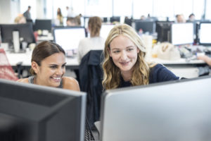 Two women working in office looking at computer, smiling. Demonstrating building a great company culture.
