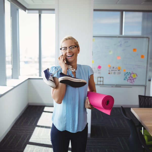 Corporate health and wellbeing - Smiling executive talking on mobile phone while holding exercise mat and shoes